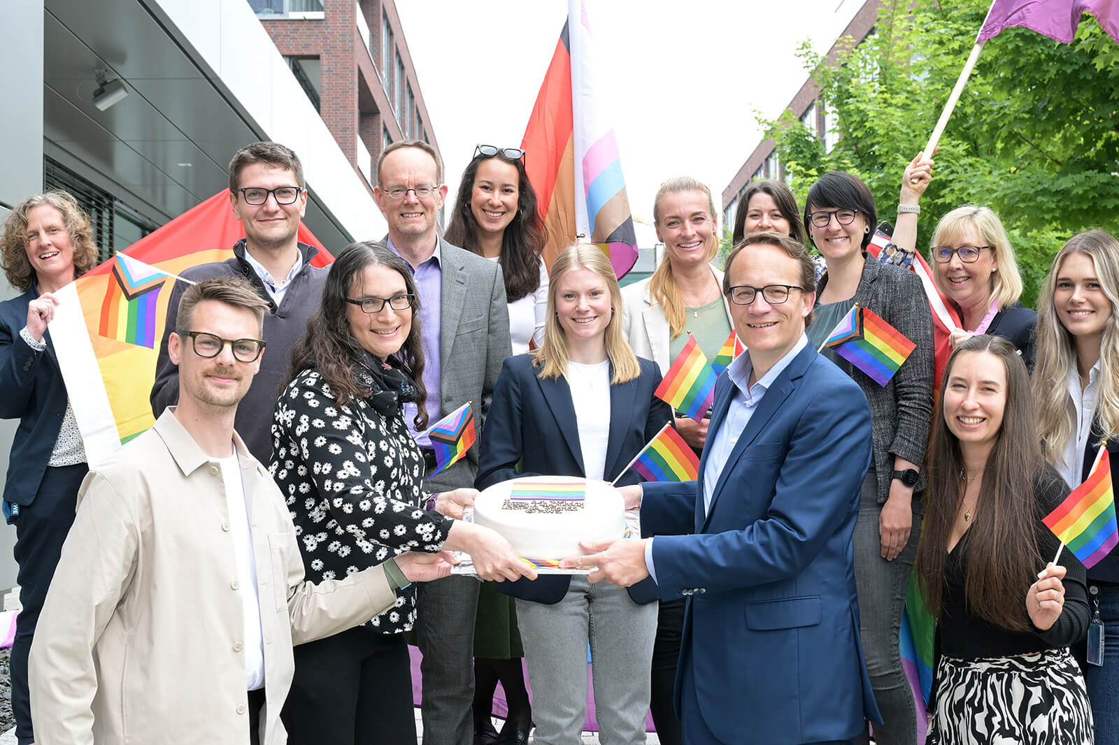 Eine Gruppe von Menschen feiert mit Regenbogenflaggen und einer Torte, um Inklusivität und Vielfalt zu fördern.