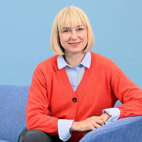 Eine Person mit kurzem blonden Bob sitzt auf einem blauen Sofa, trägt ein rotes Cardigan und ein hellblaues Hemd.