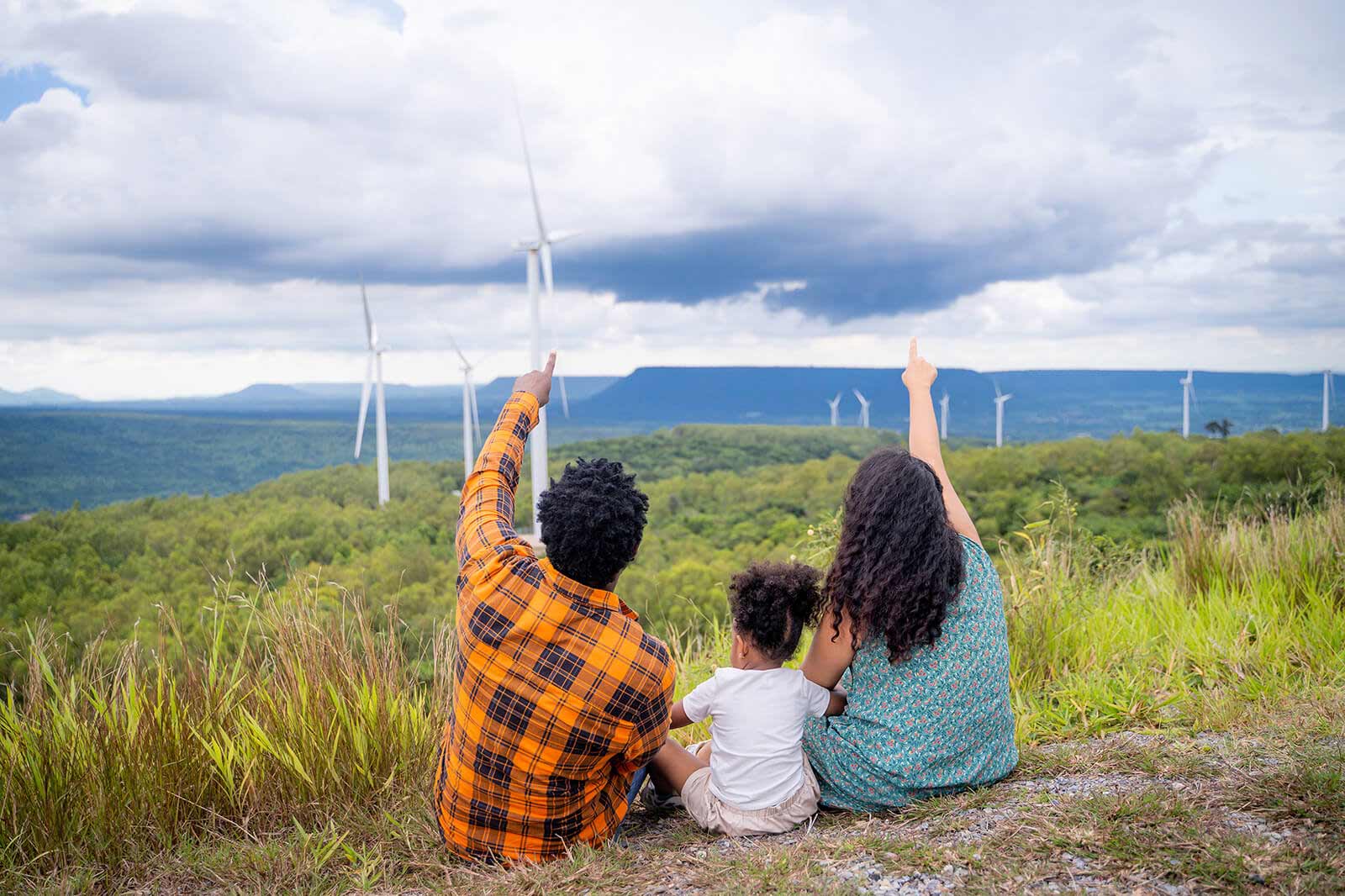 A family sits on a grassy hill, pointing at wind turbines in the distance, surrounded by lush greenery under a cloudy sky.
