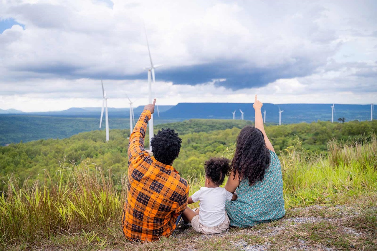 A family sits on a grassy hill, pointing at wind turbines in the distance, surrounded by lush greenery under a cloudy sky.