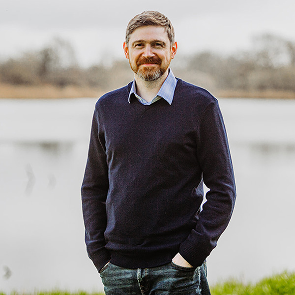 A man stands by the water's edge on a cloudy day, dressed in a dark blue jumper.