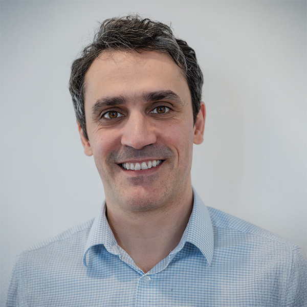 A man with short grey hair wears a checkered shirt and stands against a light wall.