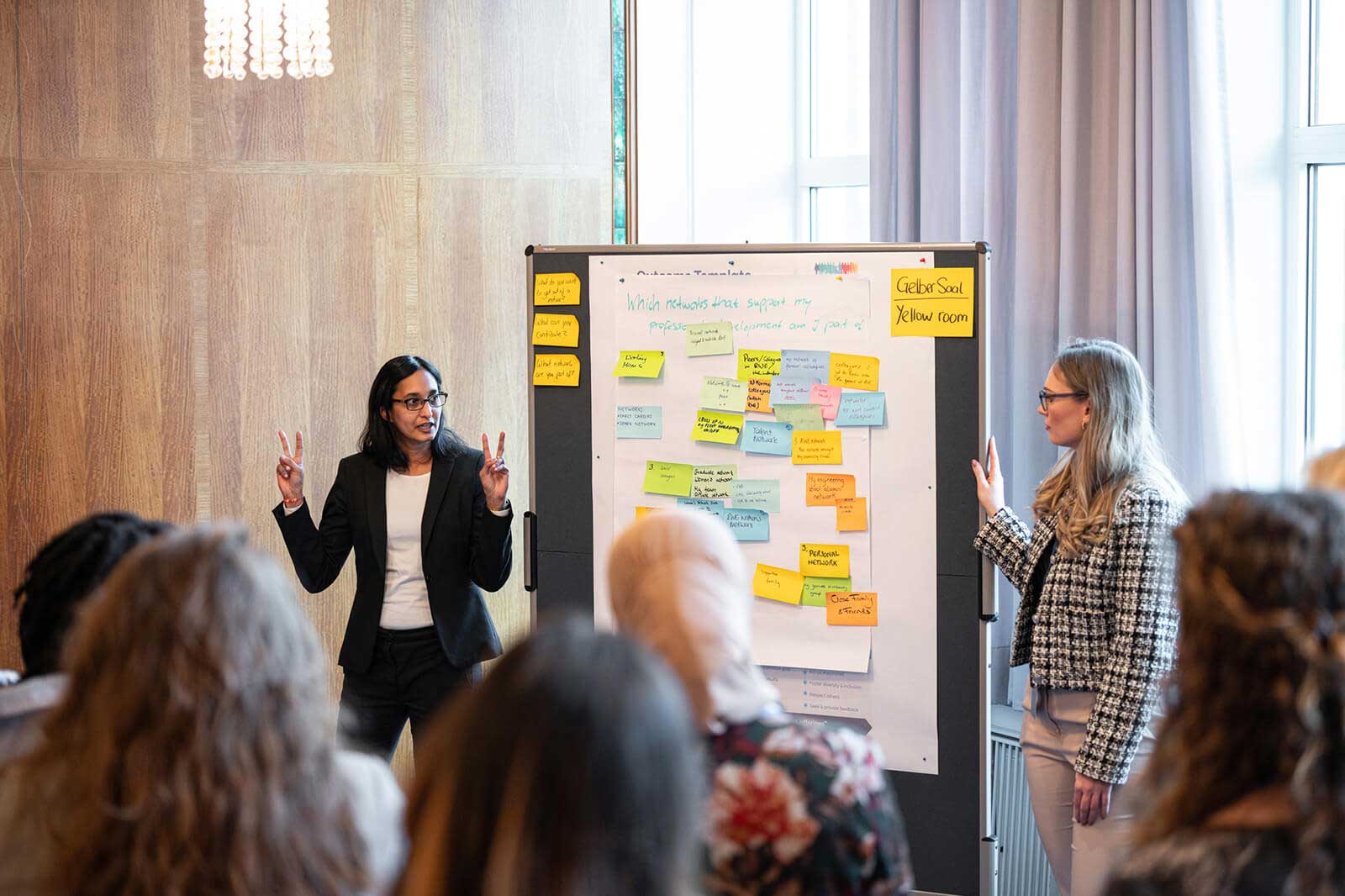 Two women present in a meeting room, standing in front of a poster board covered in sticky notes.