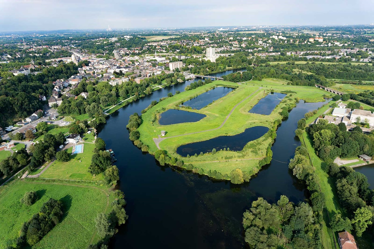 An aerial view of a city featuring green fields, water bodies, and a mix of residential buildings and trees.