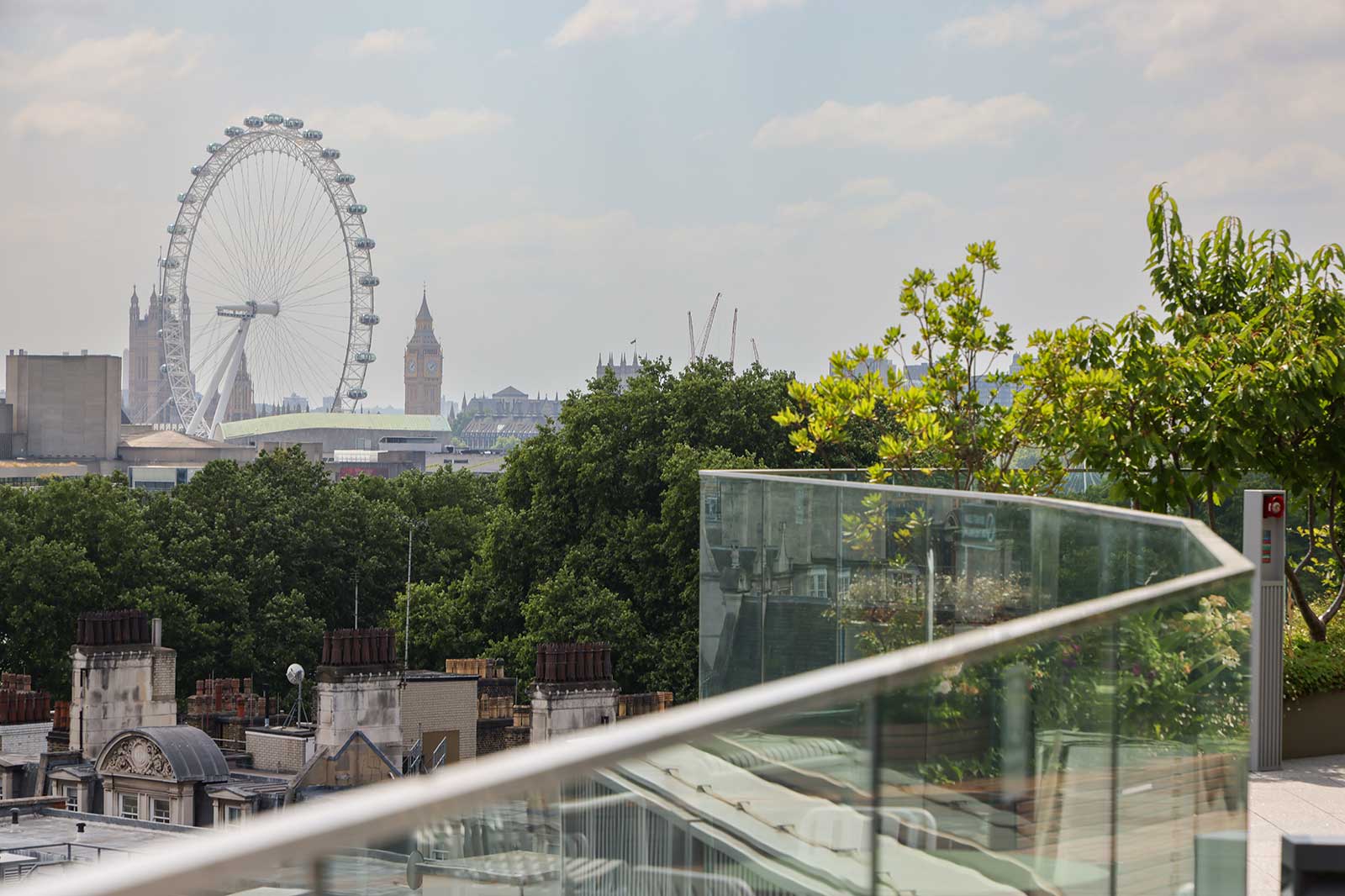 A glass balcony railing and rooftop trees foreground a tree-lined London skyline with the London Eye and Elizabeth Tower against a pale sky.
