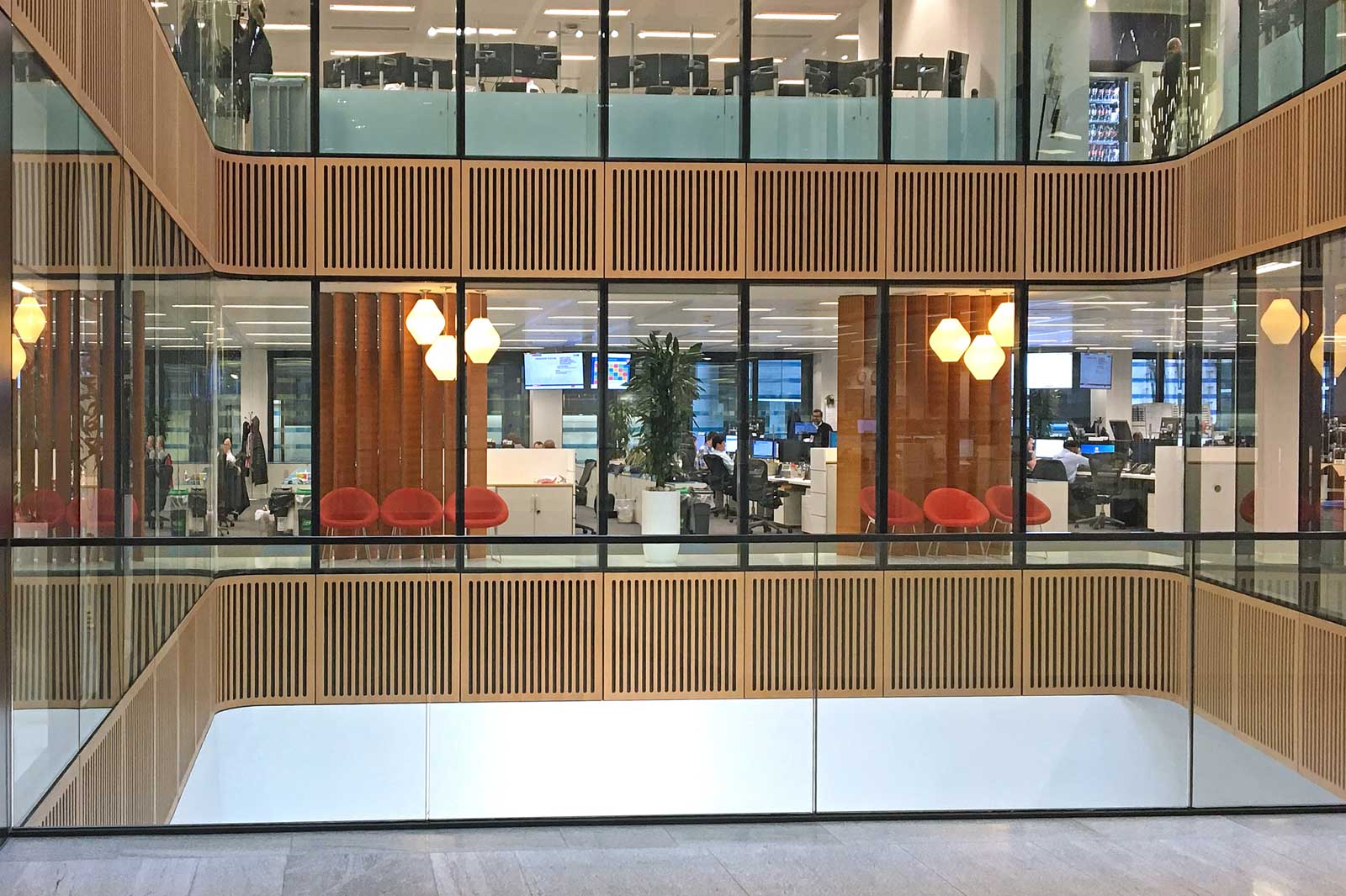 A modern office building with large windows, wooden elements, red-orange chairs, and several workstations visible.