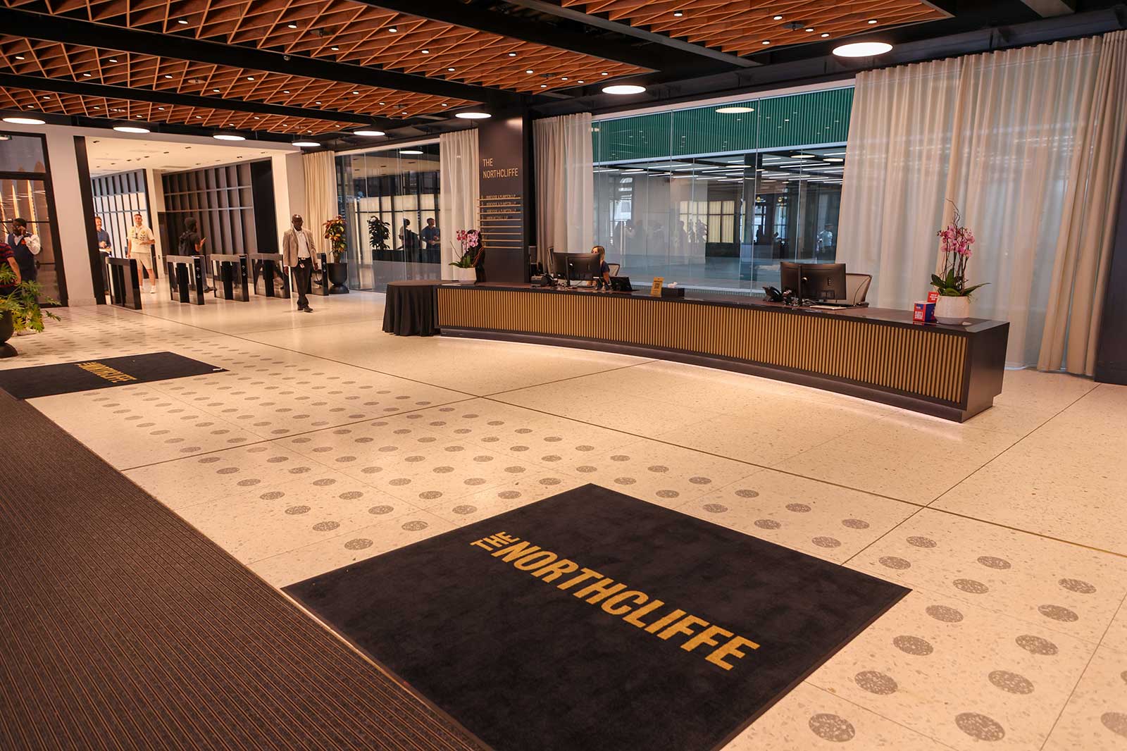 A black floor mat reading "THE NORTHCLIFFE" sits on a cream terrazzo floor before a long wooden reception desk and warm wood ceiling.