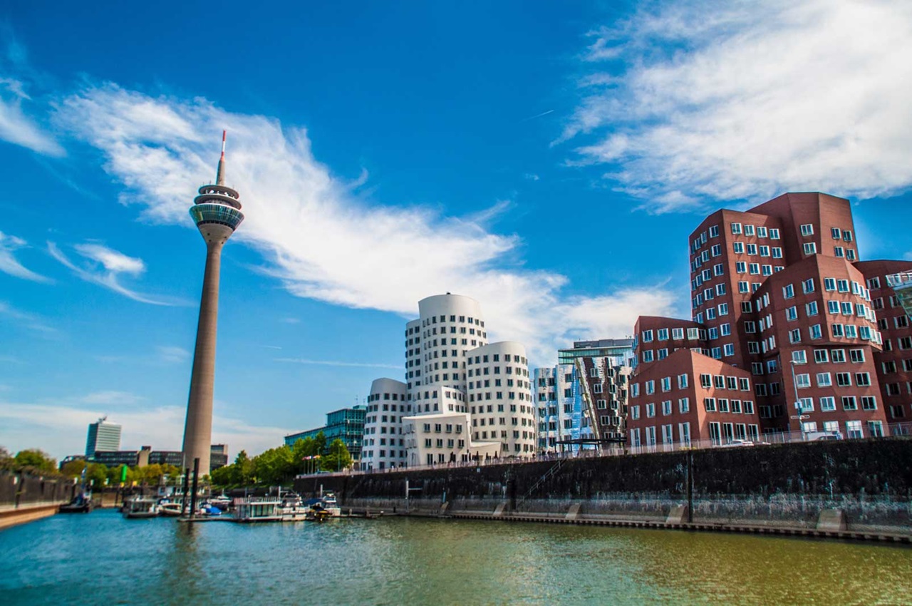 Blick auf moderne Gebäude am Wasser mit dem Rheinturm im Hintergrund und klarem Himmel.