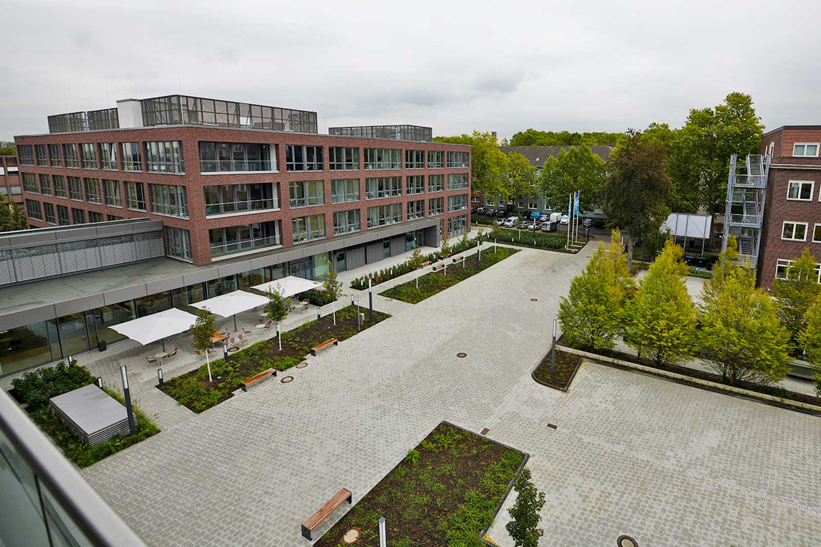 Ein modernes Gebäude mit roter Ziegelfassade und großen Fenstern umgeben von Bäumen und einer gepflasterten Fläche.