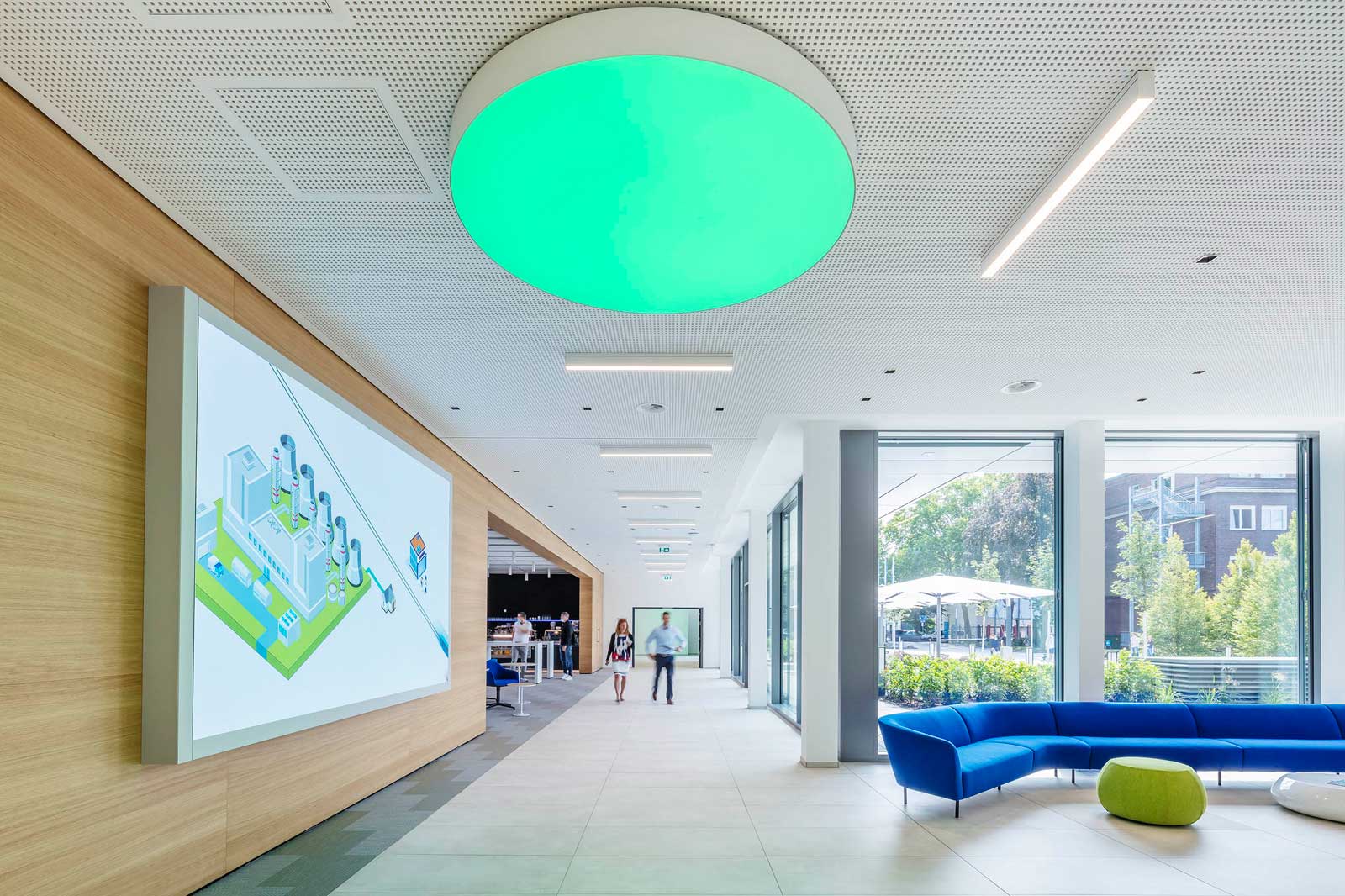 A modern interior features a green ceiling light, a large screen, and blue sofas. People walk through the space.