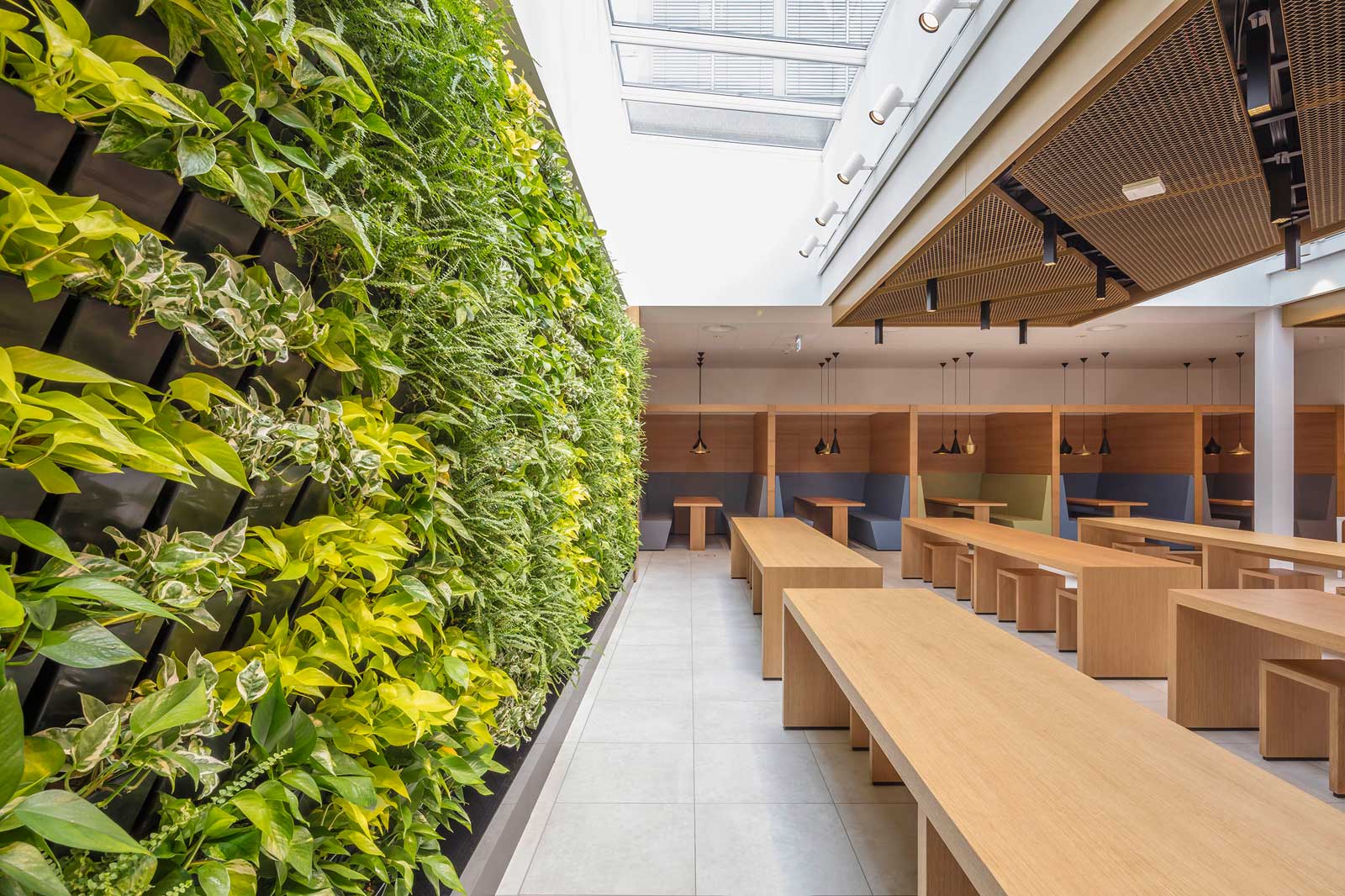 A modern room featuring wooden tables and a green wall full of plants. The space is well-lit with plenty of natural light.