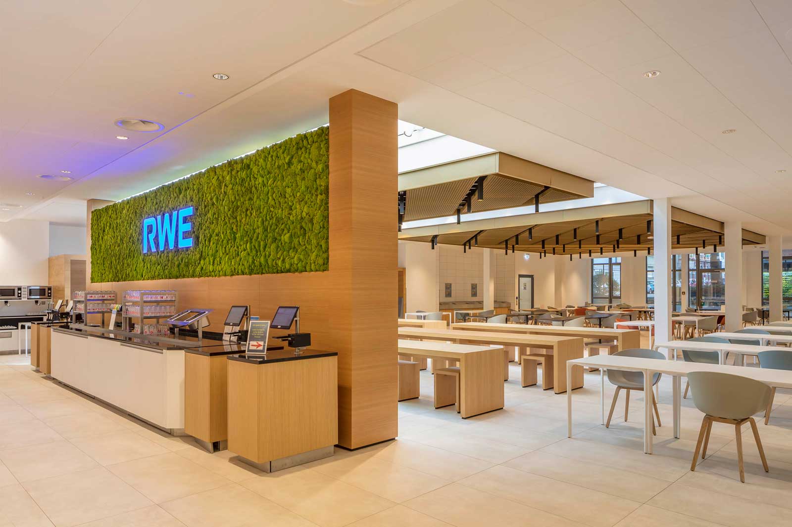 Modern cafeteria featuring a vibrant moss wall, wooden furniture, and stylish lighting. The space is bright and inviting.
