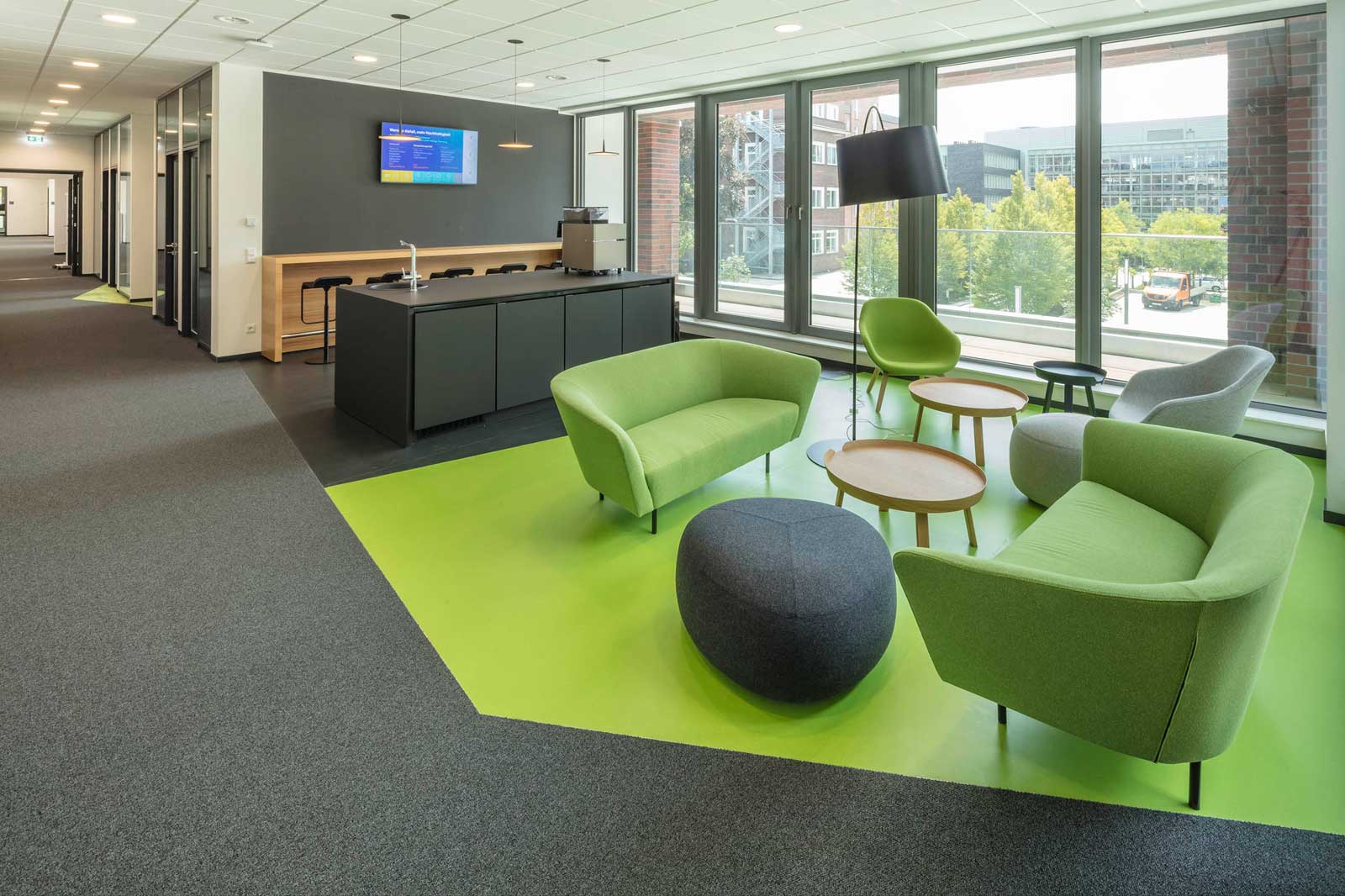 Modern office space featuring green sofas, wooden tables, and large windows. A screen and coffee machine are visible.