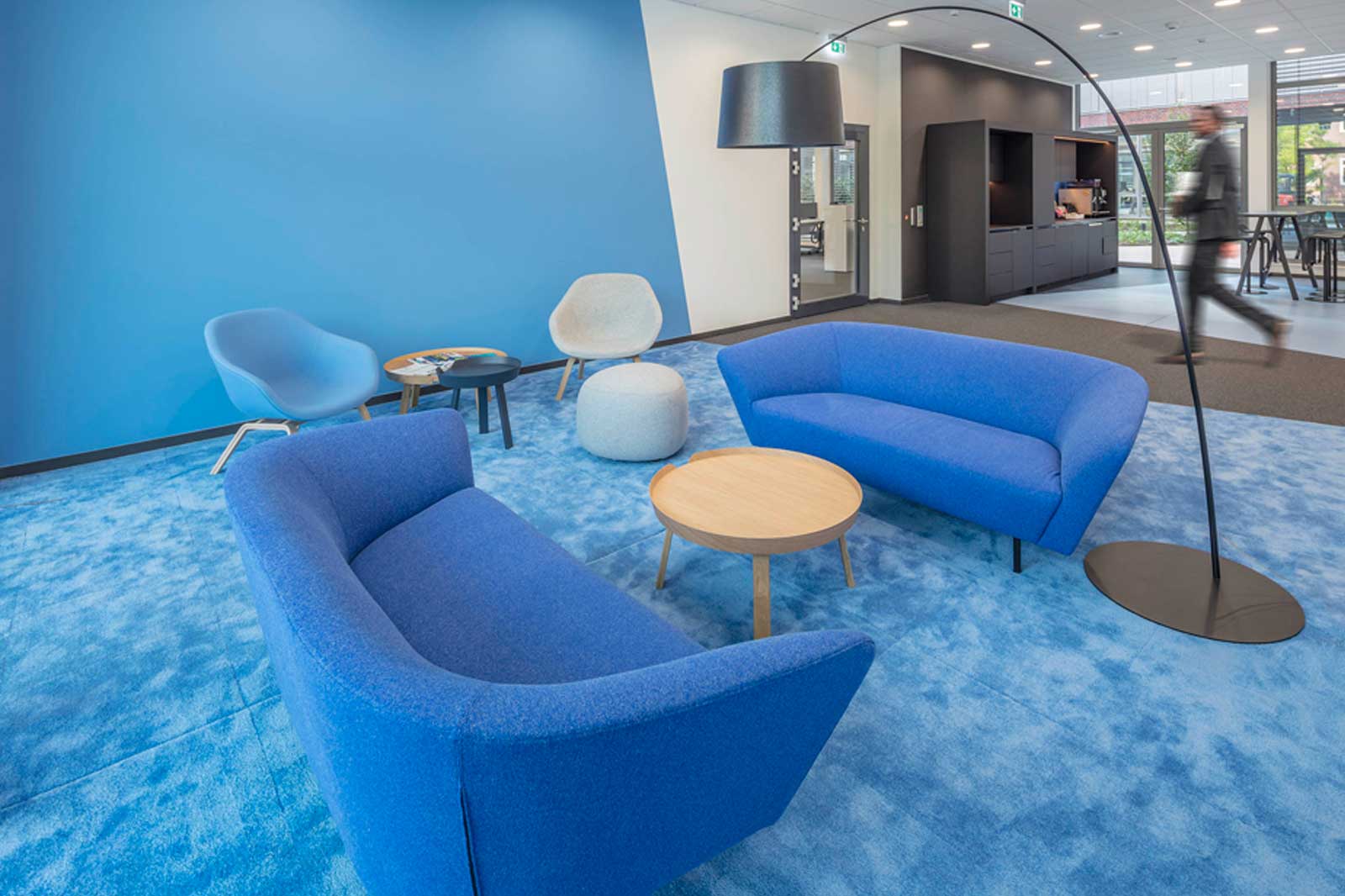 A modern lounge featuring blue sofas, a round wooden table, and a floor lamp. The floor is blue, and a visitor is walking by.