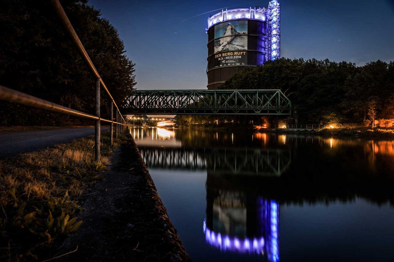 A lit industrial structure by the water at night, next to a green bridge and a paved path.