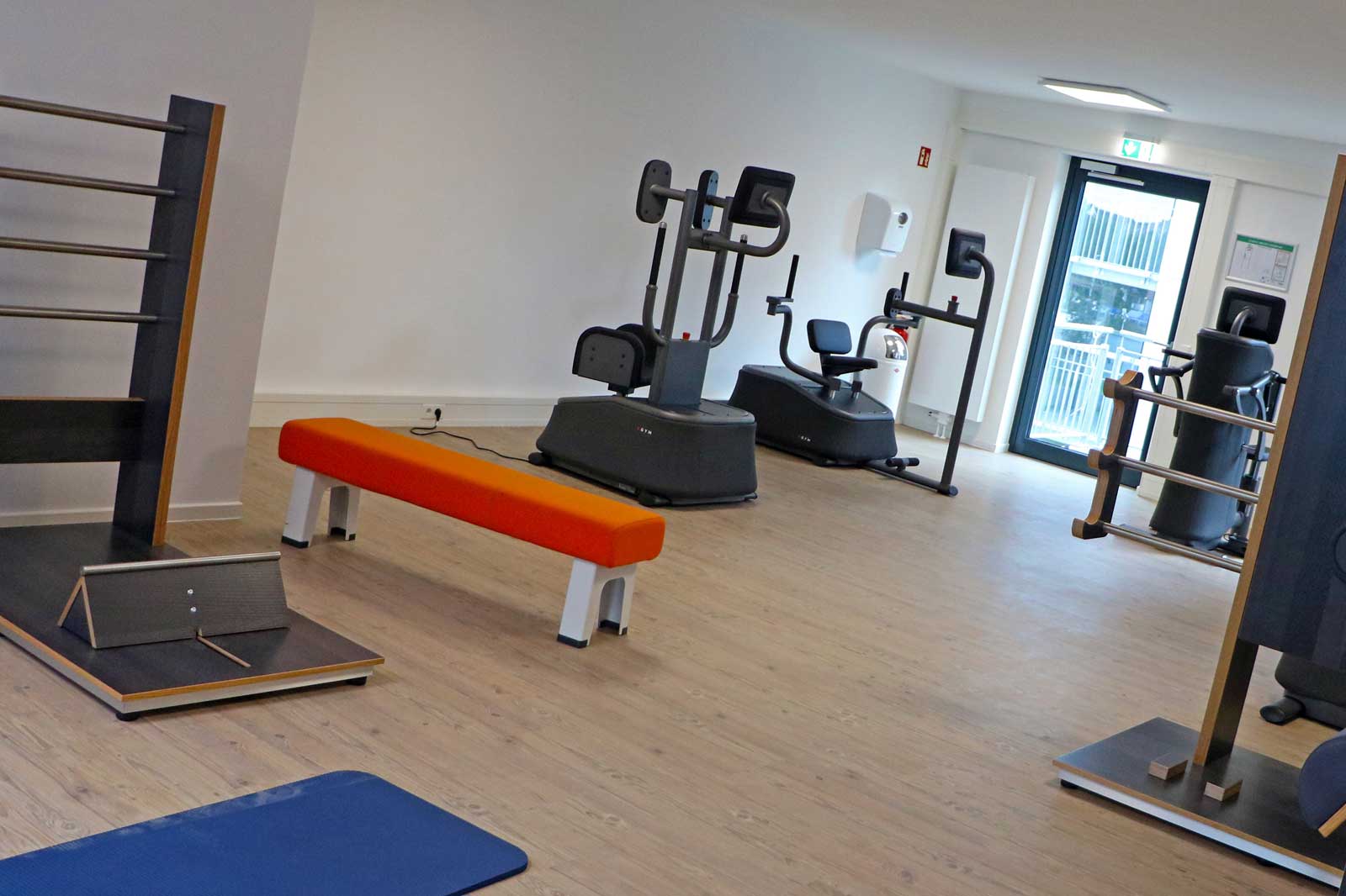 Ein modernes Fitnessstudio mit verschiedenen Trainingsgeräten, darunter eine orange Bank, Matten und ein großer Fensterbereich.