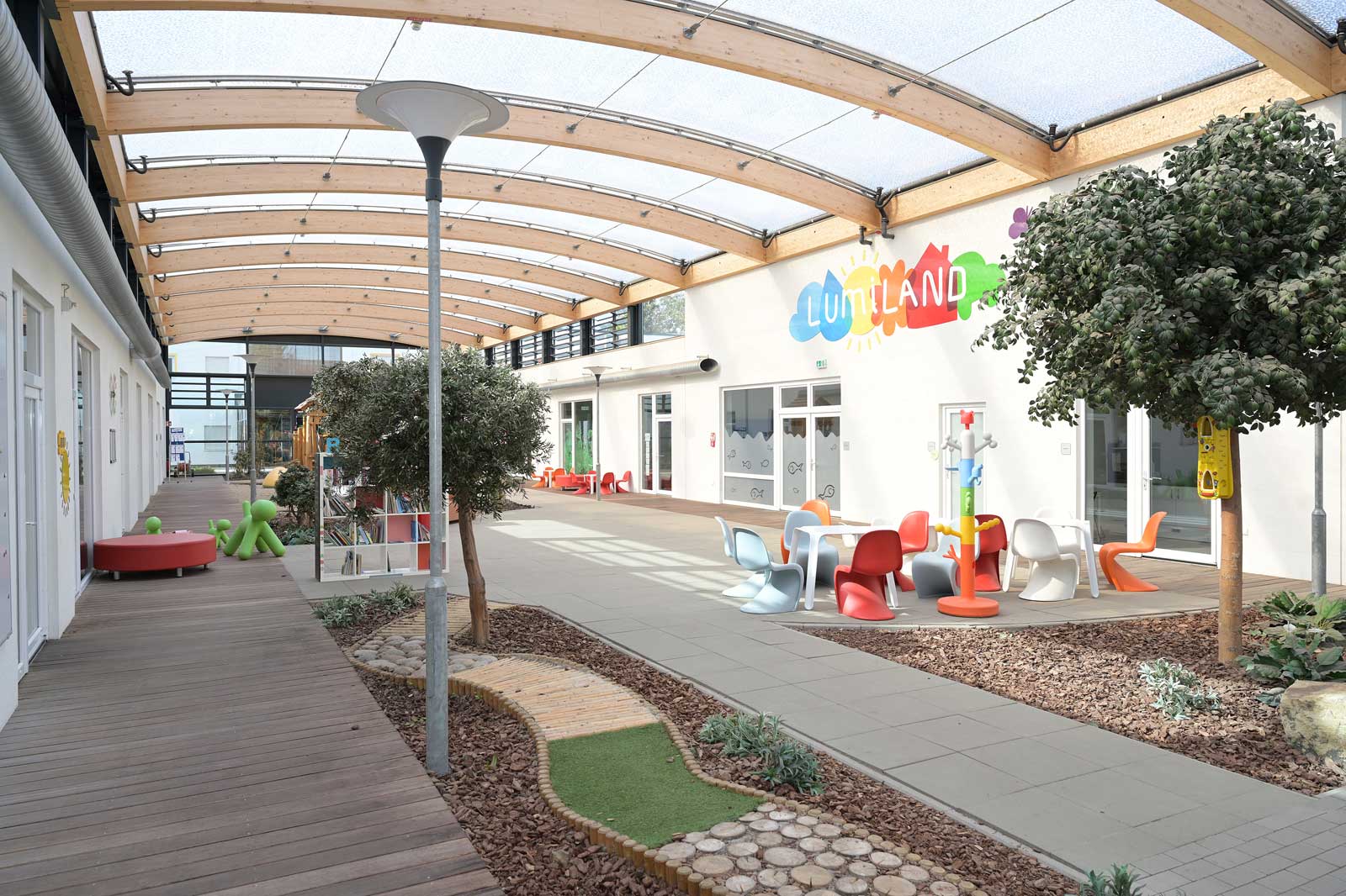 A bright courtyard featuring wooden ceilings, colourful furniture, and play equipment, surrounded by plants and vibrant wall art.