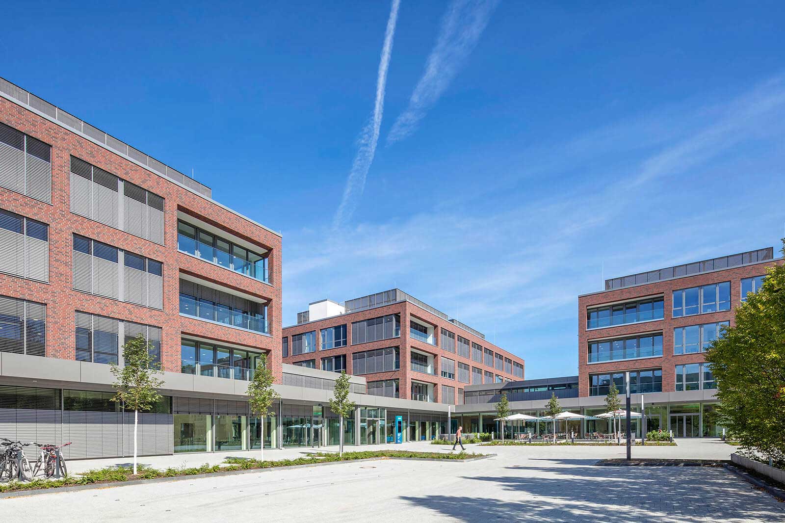 Modern red brick buildings in a well-lit courtyard with trees and bicycles under a bright blue sky.