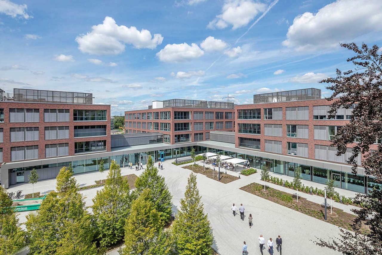 The image shows a modern brick office building with a courtyard, featuring people and trees in the scene.