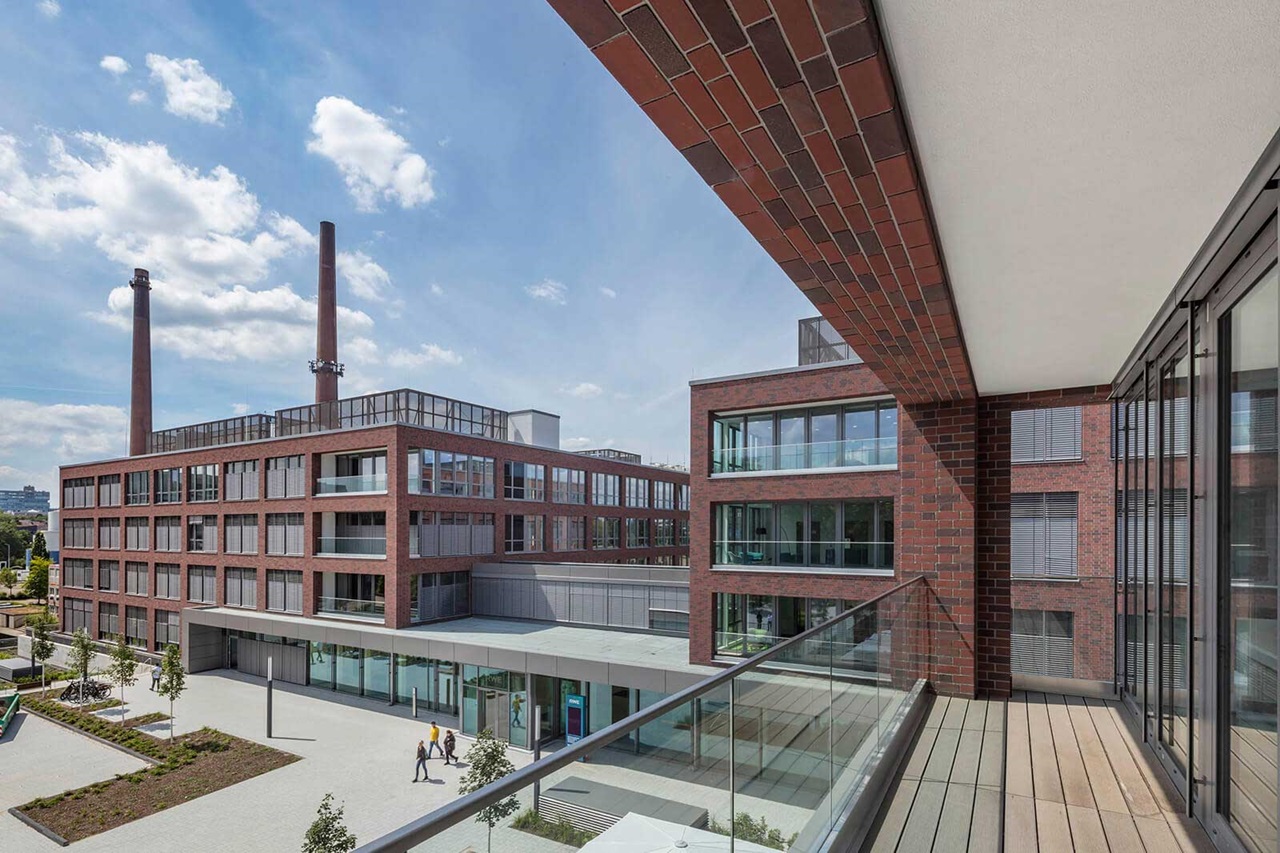 A view of modern brick buildings with large windows and smoking chimneys under a blue sky.