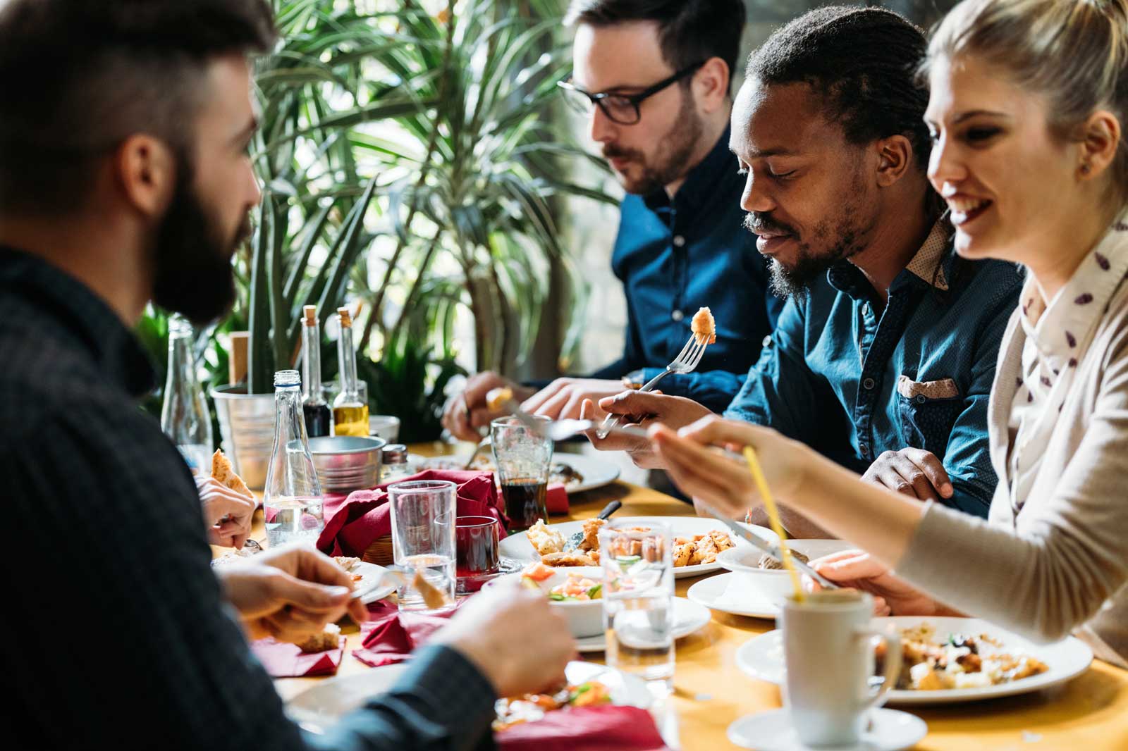 A group of people sit at a table with various dishes and drinks, surrounded by plants.