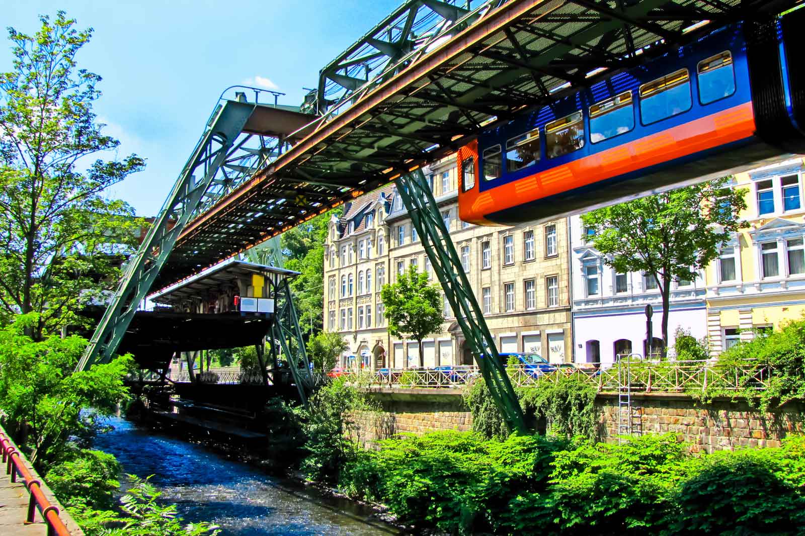 A suspended train above a small river with green banks and historical buildings in the background.