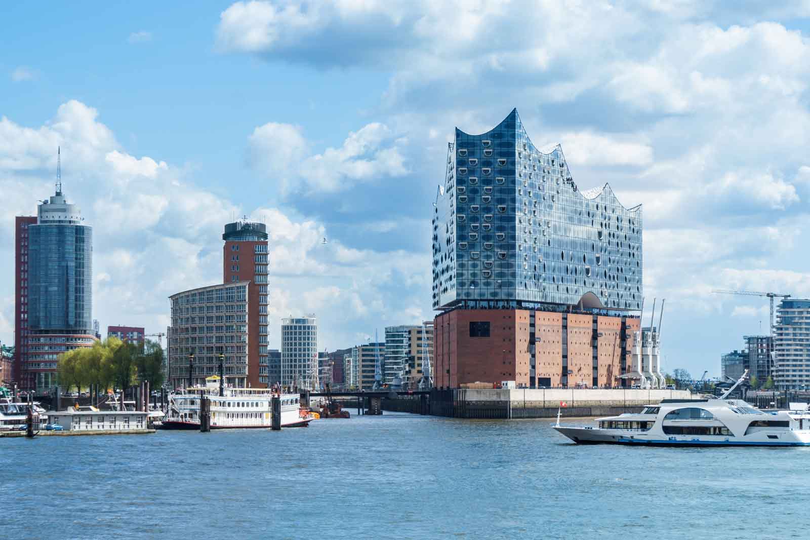 The Elbphilharmonie in Hamburg, surrounded by modern buildings and boats on the water, under a cloudy sky.
