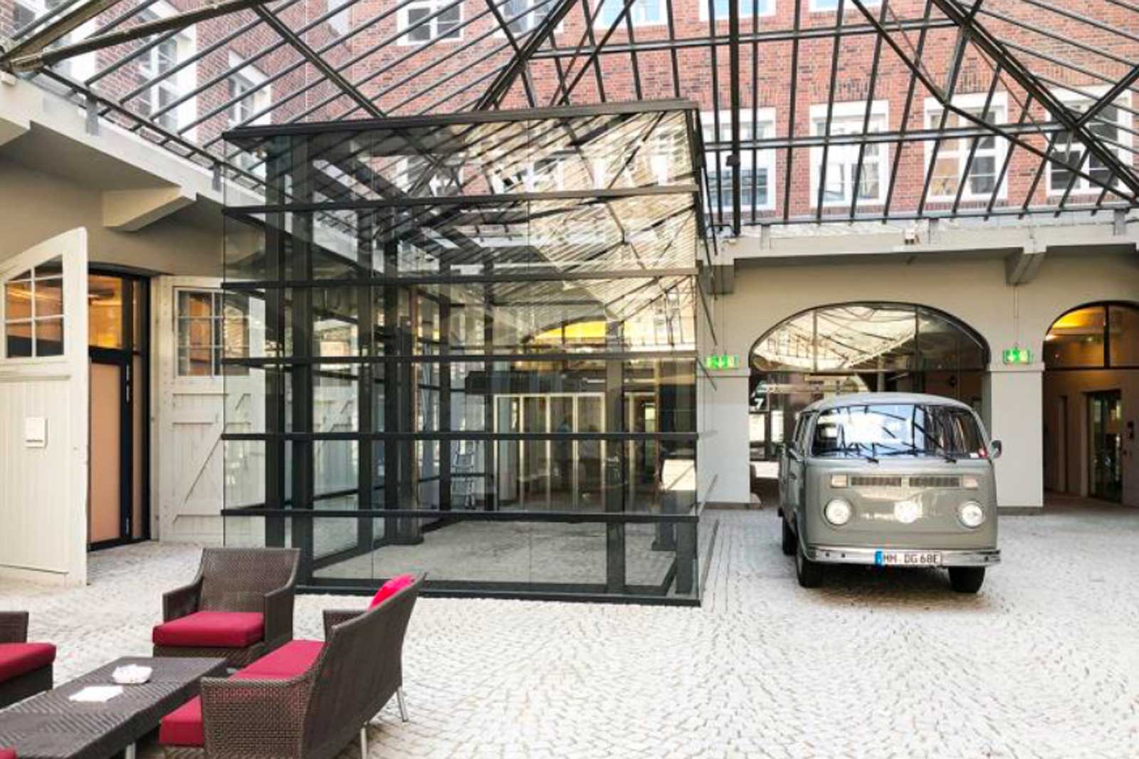 A modern glass building in a courtyard featuring an old delivery van and a seating area with red cushions.
