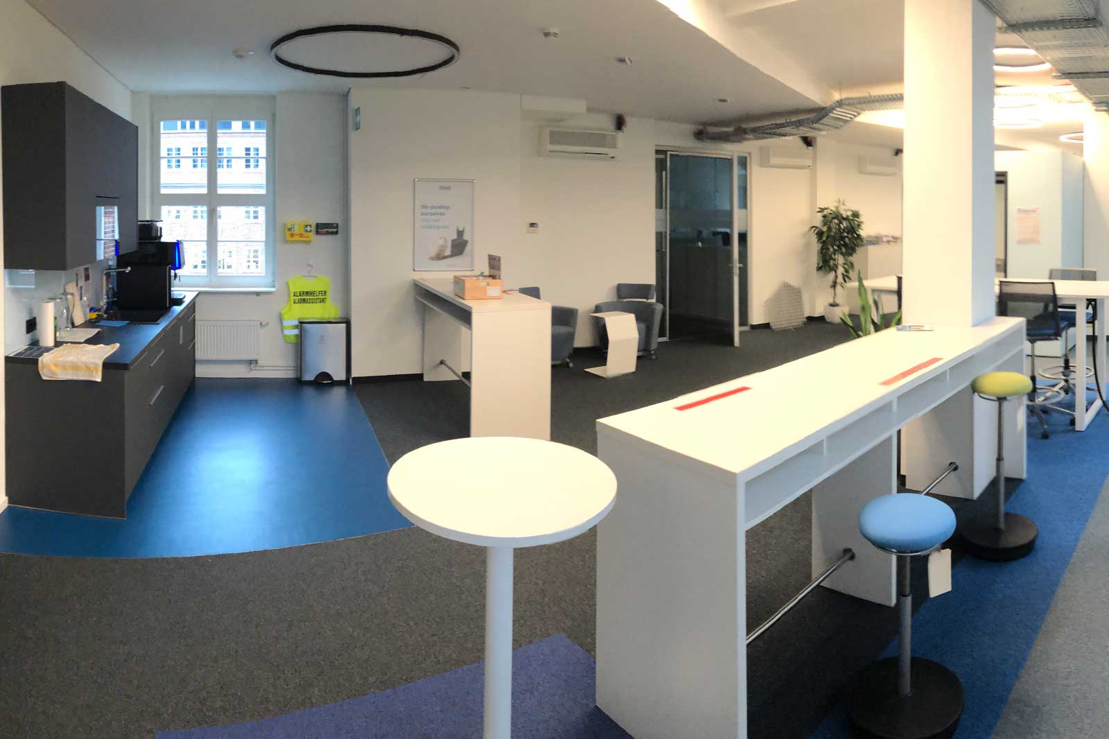 The image depicts a modern office featuring a coffee area, tables, chairs, and plants in the background.