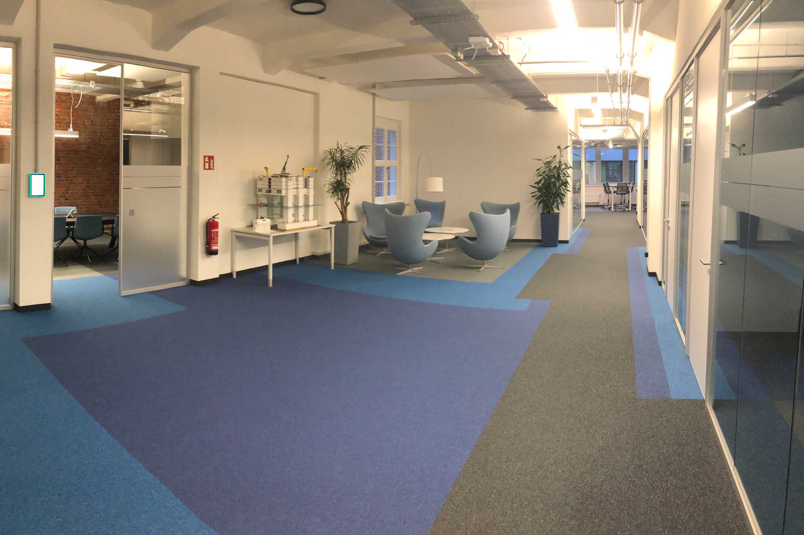 A modern office featuring blue carpets, comfortable chairs, and plants. A meeting room is visible.