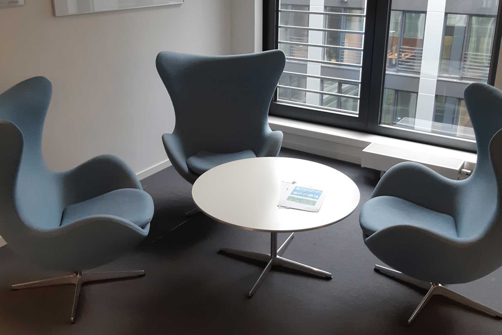 A modern seating area with three blue chairs and a round white table, featuring a tablet and large windows in a bright room.