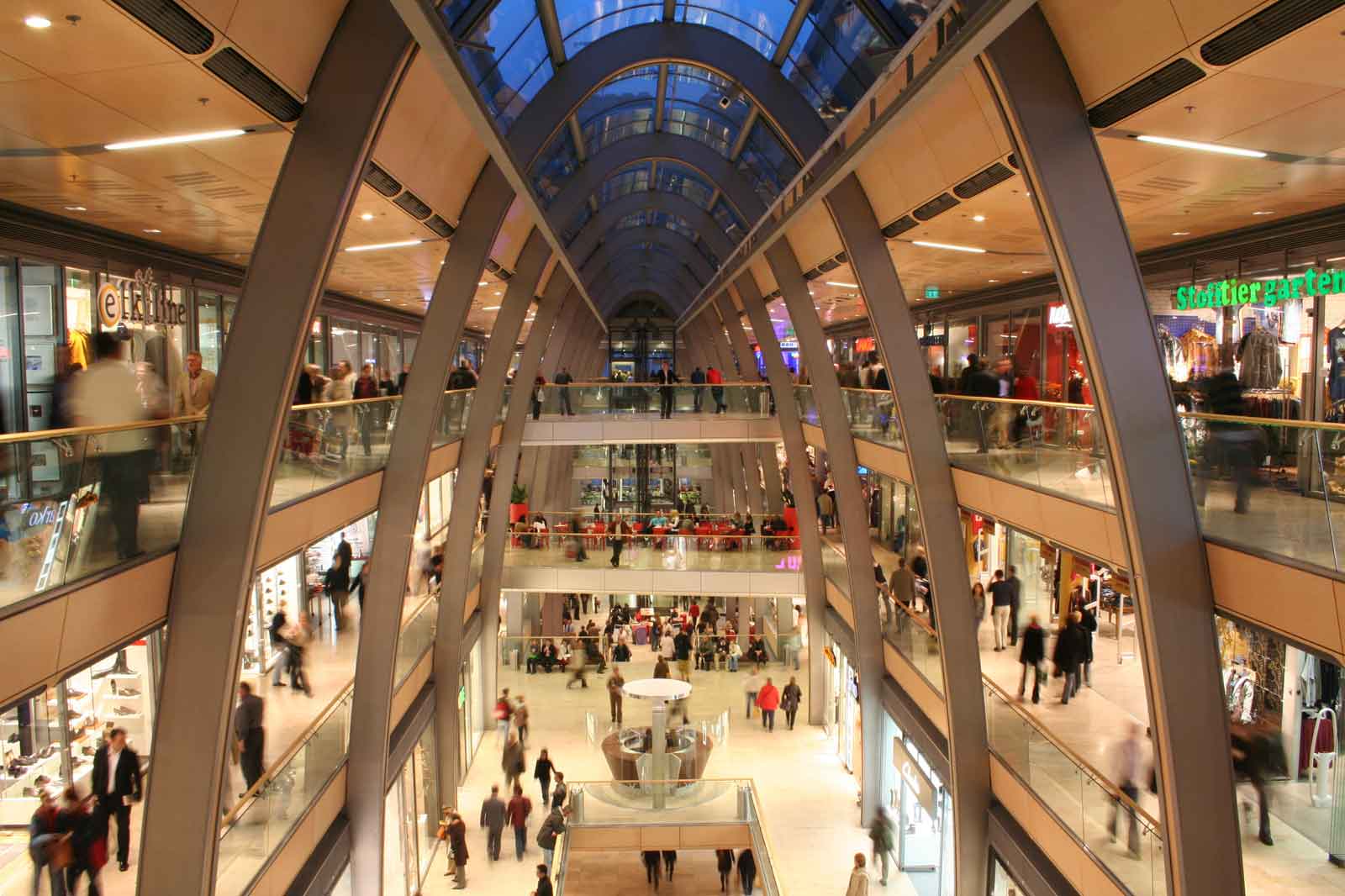 A bustling shopping centre with multiple levels, high foot traffic, and a glass roof.