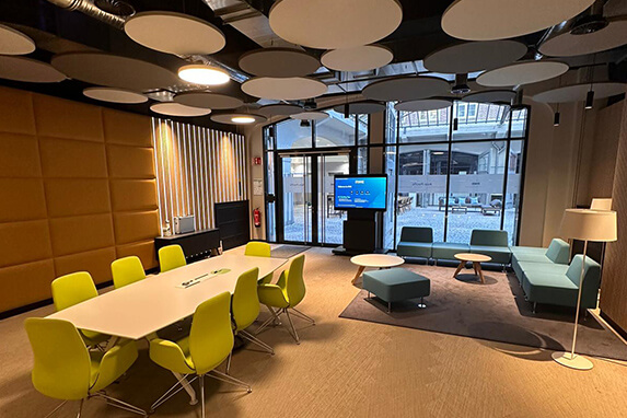 A modern meeting room featuring a large table, green chairs, a television, and a view of an indoor courtyard.