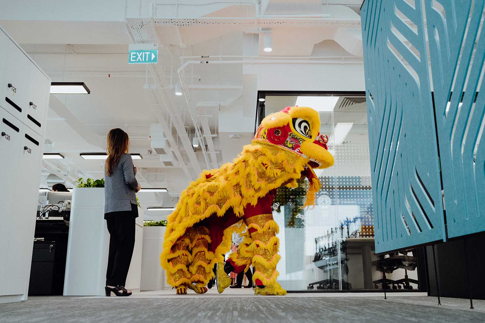 A lion dance costume in a modern office, with a person watching. The costume is yellow with colourful decorations.