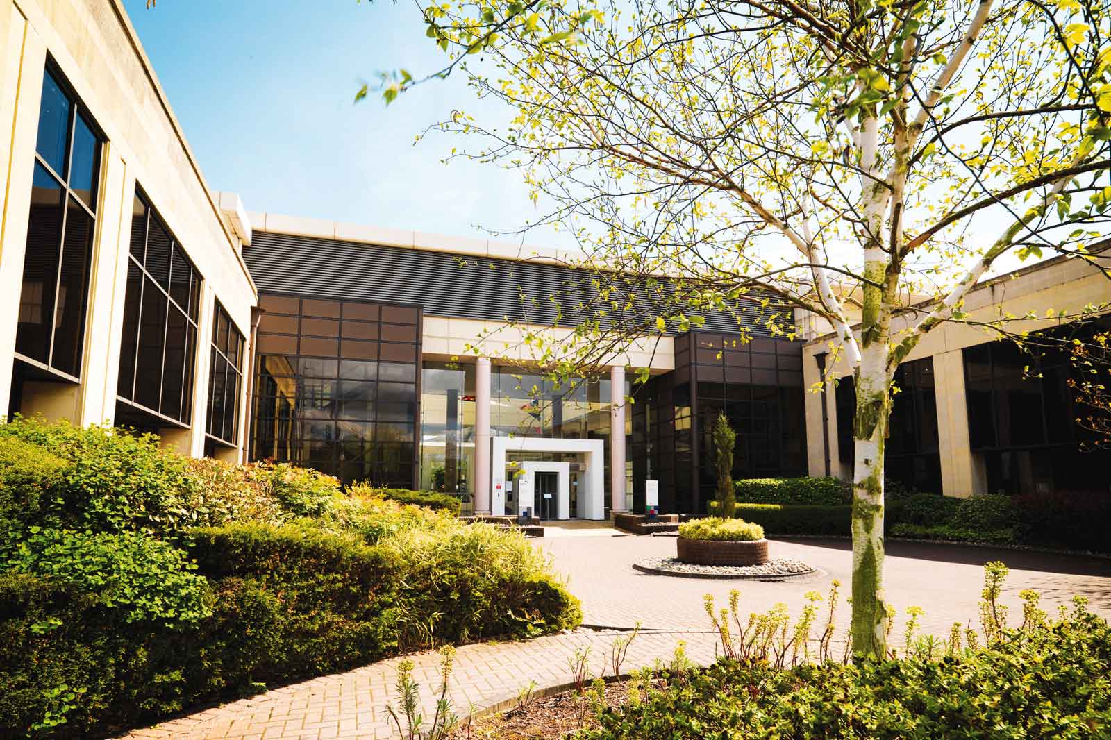 A modern building with large windows, surrounded by well-maintained gardens and trees, under a clear blue sky.