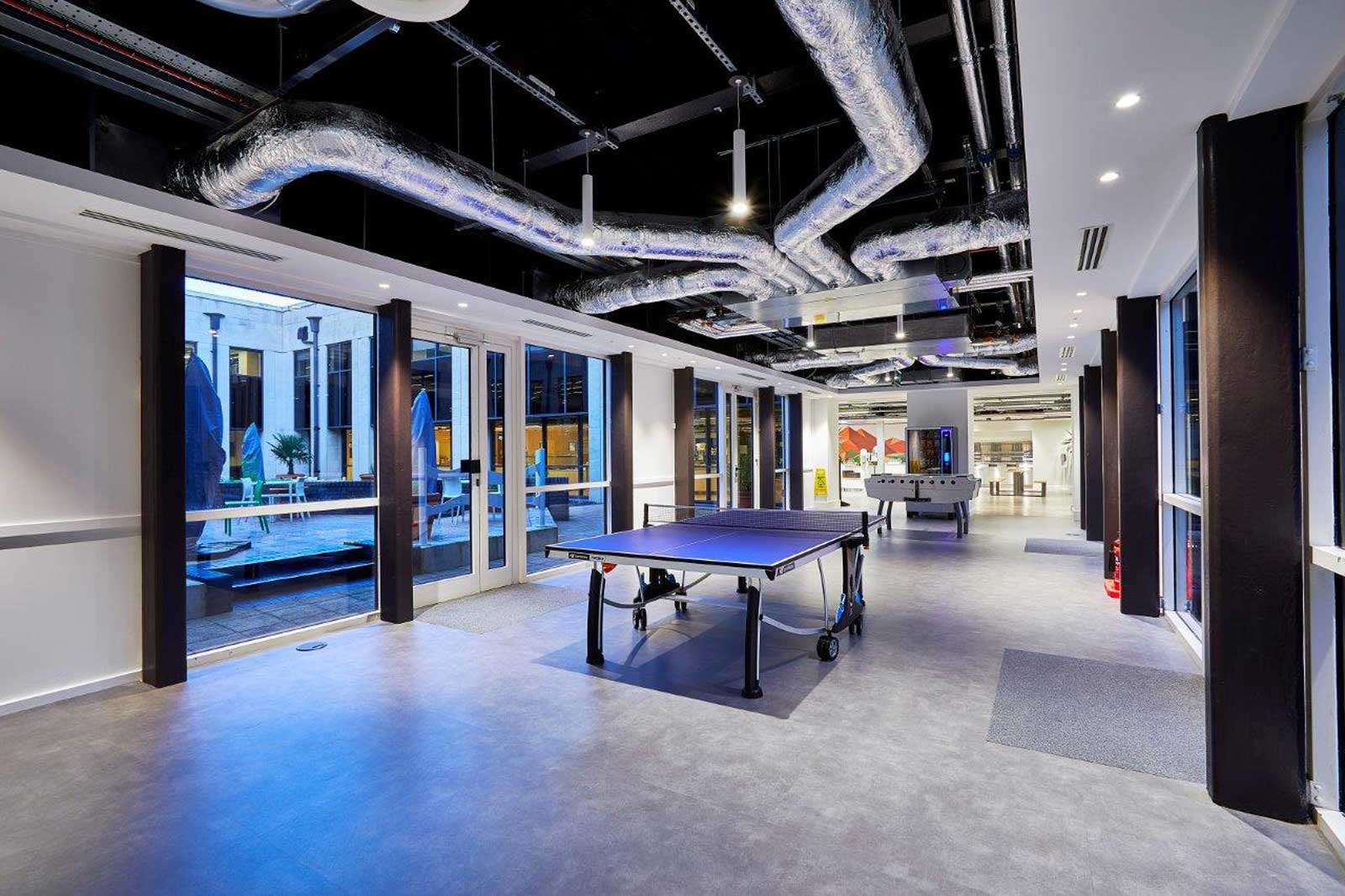 A modern room featuring a table tennis table and a foosball table, large windows, and exposed ductwork on the ceiling.