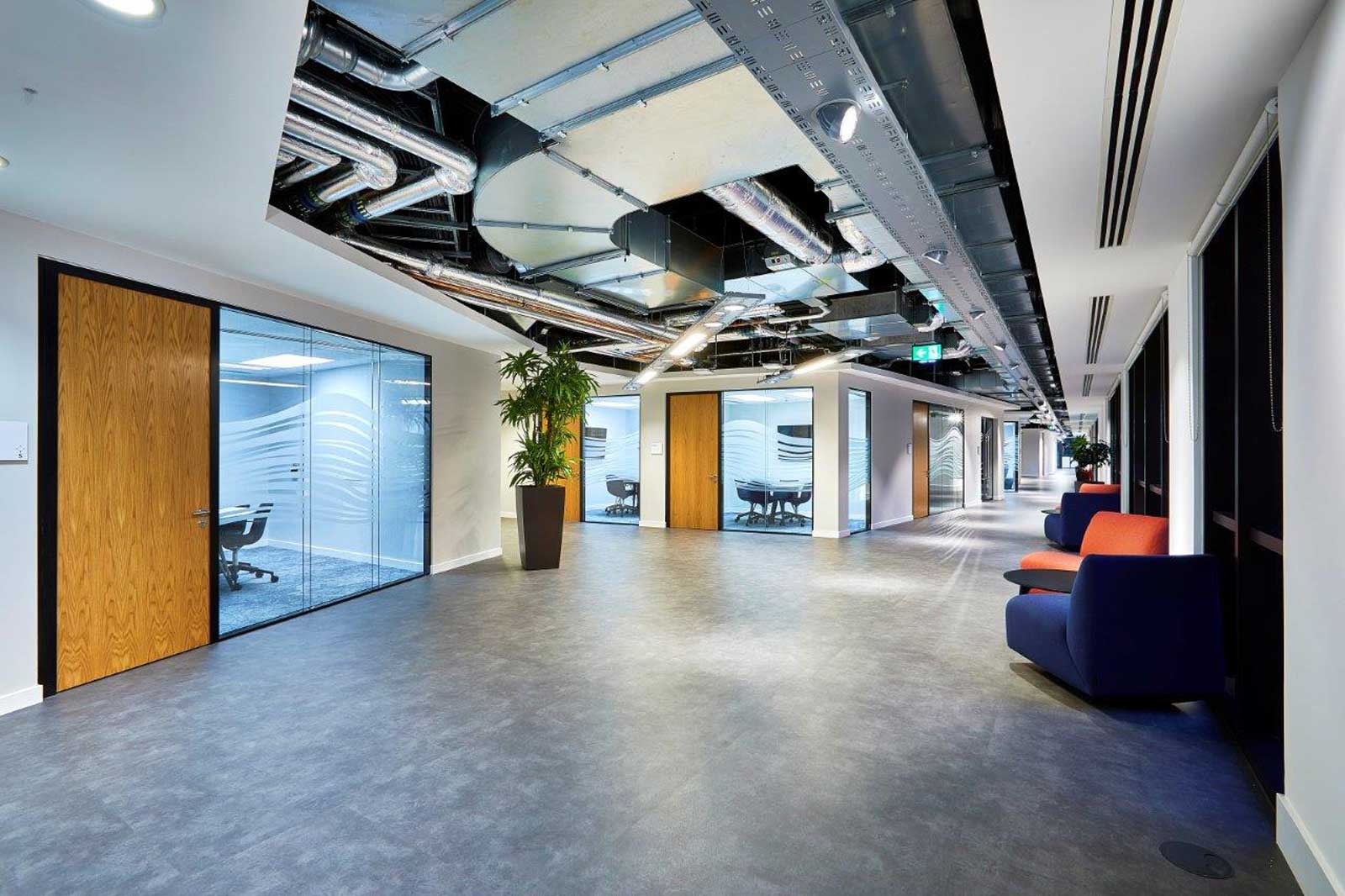 A modern office with glass doors, orange and blue seating, and a green area. The ceiling features visible pipes.