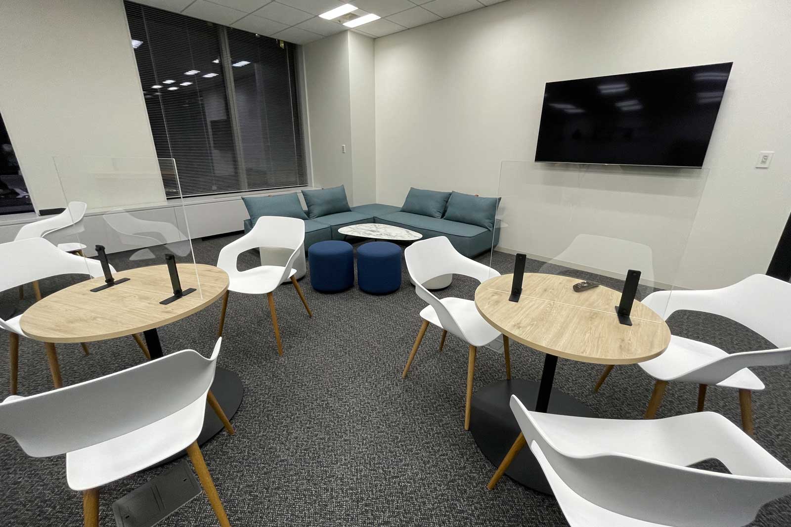 A modern meeting room with tables, chairs, dividers, and a couch. A television is mounted on the wall.