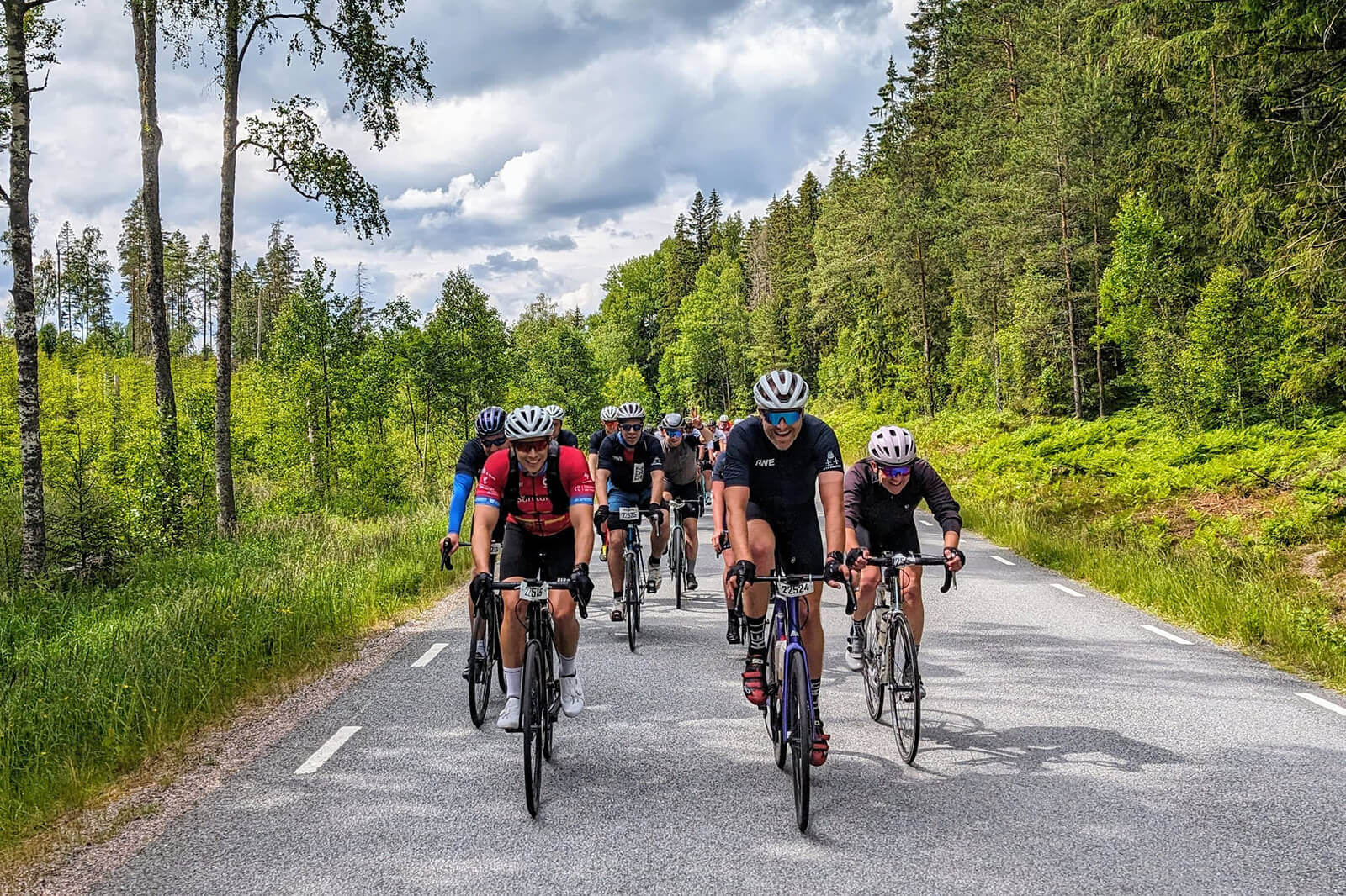 A group of cyclists riding on a rural road, surrounded by green trees and a cloudy sky.