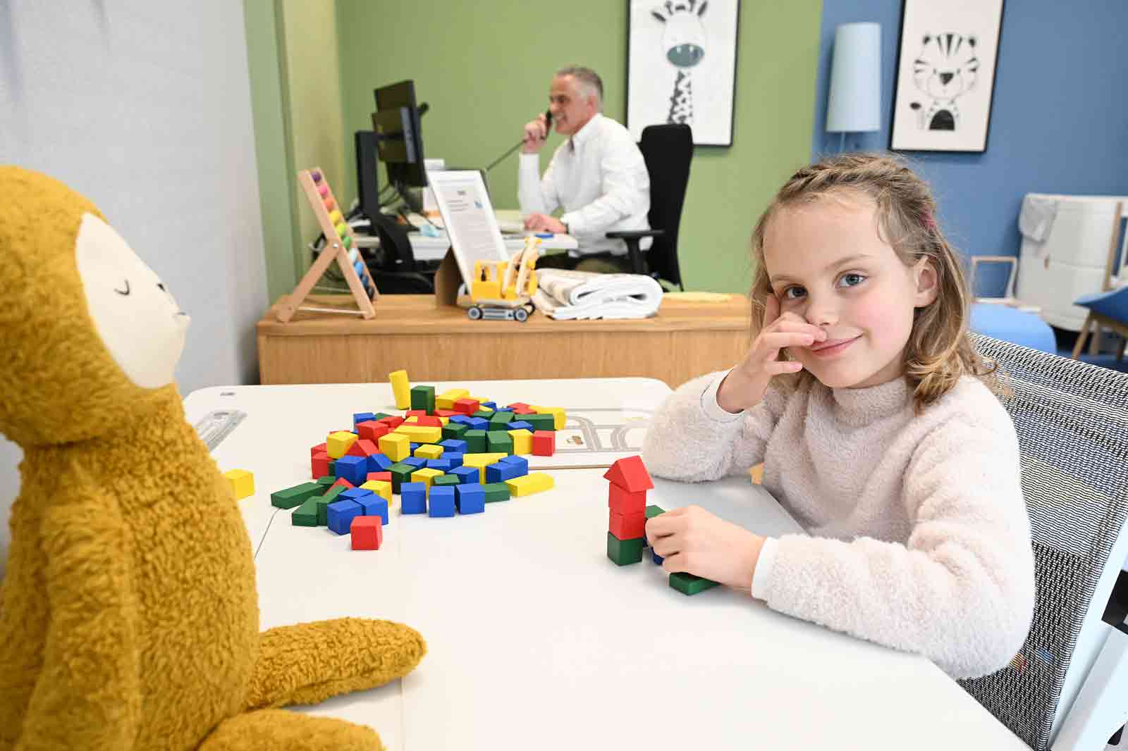 A young child builds with colourful blocks on a table, with a plush toy and a blurred adult in the background.