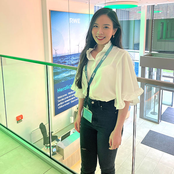 A woman stands in a modern office wearing a ruffled white shirt and black jeans. There is a reception desk in the background.