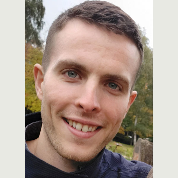 A person with a blurred face, wearing a dark top, poses outdoors among trees displaying autumn foliage.